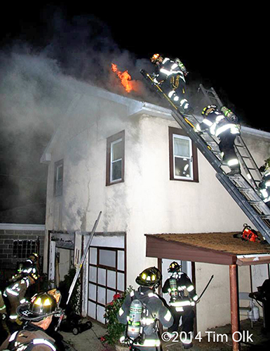 firemen climb a ladder to the roof of a house on fire