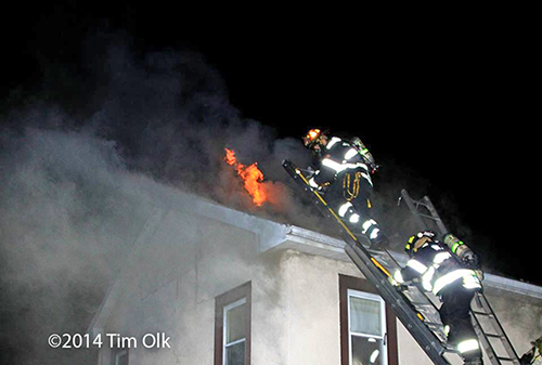 firemen climb a ladder to the roof of a house on fire