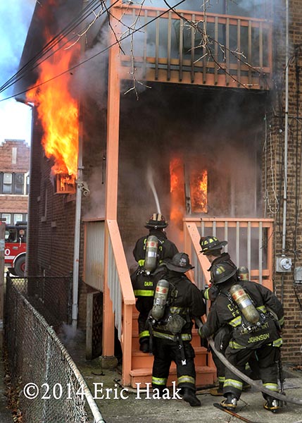 firemen with hose enter house with flames