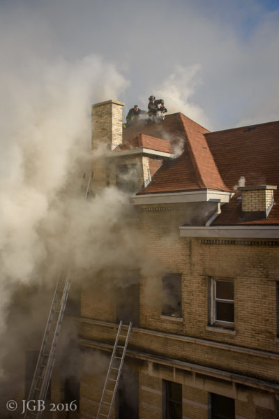 firefighters venting a roof as smoke billows from large home